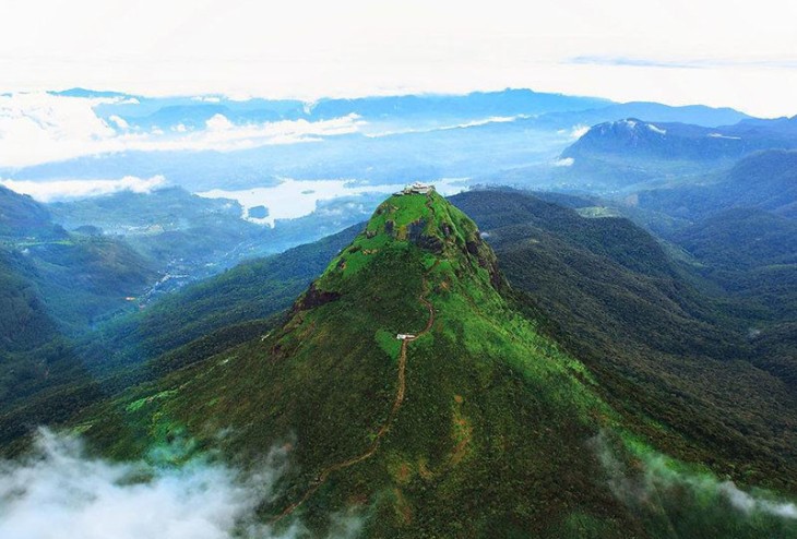 Aerial view of a forested conical mountain peak with a small summit building, rising above misty hills.